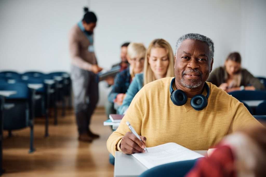 Black senior student writing a test during adult educational training course and looking at camera.